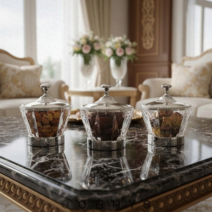Three decorative glass containers on a marble table with a blurred background of a living room. Sobhys Crystal-Cut Acrylic Bowl with Silver-Tone Metal Lid – Side Profile View
