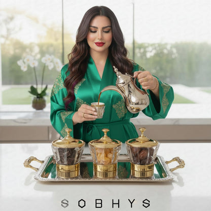 Woman in a green robe pouring coffee into a cup with a silver teapot, surrounded by coffee pots on a tray.