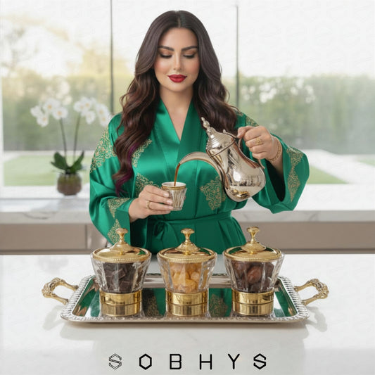Woman in a green robe pouring coffee into a cup with a silver teapot, surrounded by coffee pots on a tray.