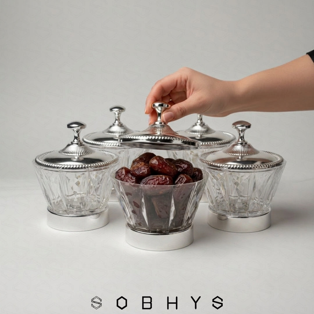 Silver canisters with glass bowls containing dates, hand reaching to open one, on a light gray background. Sobhys Luxury Serving Bowl – Silver-Tone Metal Base on Wooden Surface
