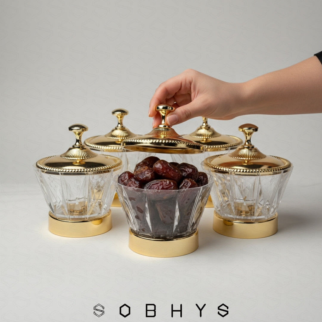 Hand reaching for a glass container with gold lid filled with dates, on a light gray background with 'SOBHYS' branding. Sobhys Luxury Serving Bowl – Gold-Tone Metal Base on Wooden Surface