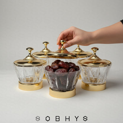 Hand reaching for a glass container with gold lid filled with dates, on a light gray background with 'SOBHYS' branding. Sobhys Luxury Serving Bowl – Gold-Tone Metal Base on Wooden Surface