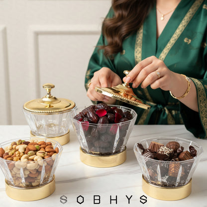 Person in a green robe using a gold container to serve snacks from glass jars on a white surface, with 'SOBHYS' branding. Sobhys Gold-Tone Acrylic Bowl – Top View Showing Diamond-Cut Facets and Ornate Lid Detail